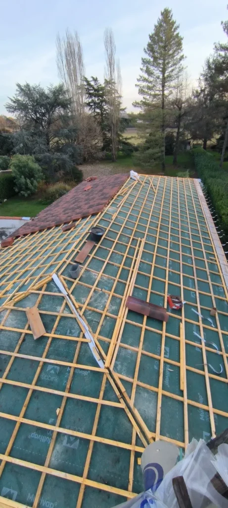 Rénovation de toiture : pose du lattis sur pare-vapeur Multivap Lattis de toiture en construction, posé sur pare-vapeur Ubbink Multivap. Tuiles brun-rouge installées sur une partie du toit, vue nature.